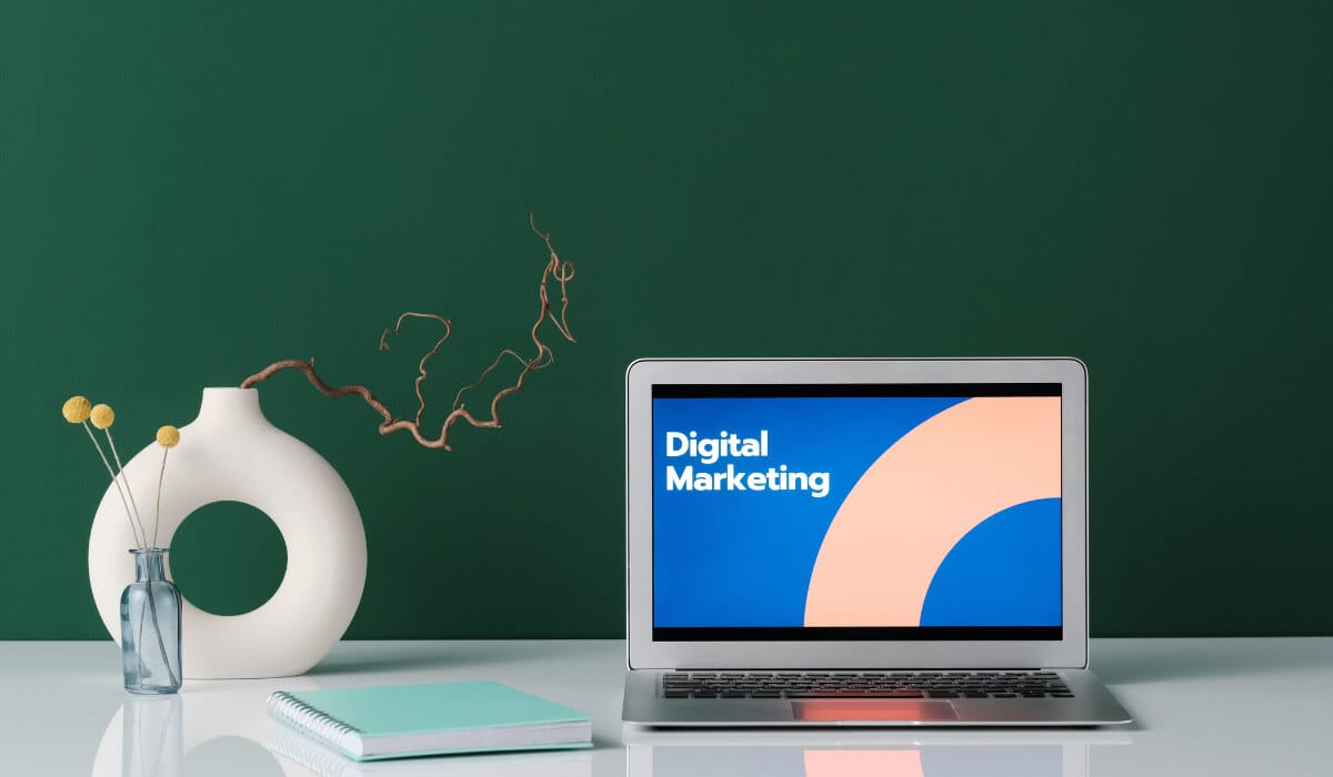 laptop on table, digital marketing written on screen, a notebook and a flower vase next to the laptop